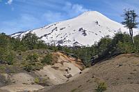 Villarrica  Villarrica steht für 'reicher Flecken', womit eigentlich nur die Schönheit und Vielfalt der Natur gemeint sein kann, denn die gleichnamige Stadt soll eher unansehnlich sein. Viel eher spielt das Städtchen Pucón als Mittelpunkt des touristischen Geschehens auf, liegt es doch viel näher am Lago Villarrica und am Volcán Villarrica. Dieser Vulkan ist bereits sehr alt, die große Caldera hat sich bereits im Pleistozän gebildet. Vor ca. 3500 Jahren öffnete sich der noch heute aktive Ausbruchkrater mit ca. 2 km Durchmesser. Seit dem sechzehnten Jahrhundert finden Aufzeichnungen über Aktivitäten statt. Danach ist es 1575, 1640 und 1948 zu schweren Ausbrüchen gekommen, während 1971 eine durch Lava gebildete Eis- und Schlammlawine diverse Häuser, Straßen und Brücken zerstörte.  Der 2840 m hohe Vulkan hat auf seinen Flanken Nebenkegel und Fissuren, die aber seiner ebenmäßigen Form nicht viel anhaben. Ständig steigt eine leichte Rauchfahne aus dem Krater auf, die die touristische Eroberung der Kraterkante durch geführte Touren aber nicht behindert, einige Skilifte zeugen außerdem von einem recht entspannten Verhältnis zwischen Vulkan und Anwohnern.  Wie seine 'Nachbar-Bilderbuchvulkane' Llaima und Osorno überrascht auch der Volcán Villarrica mit einer nur 'mittelprächtigen' Höhe von unter 3000 m. Dies erstaunt deswegen, weil die auch im Sommer weitgehend schneebedeckten Kegel eine erheblich größere Höhe vermuten lassen – besonders im Vergleich zu den oft kahlen Sechstausendern im Norden Chiles. Im Norden tummeln sich die einer Hochebene entsteigenden Vulkane aber im tropischen 15°-20° Breitengrad. Die drei Schneegrazien des Seengebiets liegen schon erheblich kälter zwischen dem 38°-40° Breitengrad. Das Klima und der Bewuchs Mittelchiles weisen daher auch viele Ähnlichkeiten zu europäischen Standards auf, was den kolonialistischen Zulauf in diese Breiten erklärt.