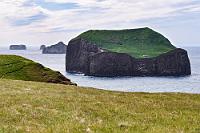 Alternativprogramm  Der Sturm bleibt uns aber erhalten, als wir die hölzerne Pforte zum Trampelpfad passieren, der um den halben Hügel herum zu den Papageitauchern führt. Schon ganz begierig auf unsere ersten Aufnahmen, führen wir unser gesamtes Equipment mit, werden aber etwas enttäuscht, da es wohl selbst den Vögeln heute zu windig ist. Von der Aussichtsplattform ist die steile Felswand, auf der Möwen und Sturmvögel brüten, weit entfernt und auf dem davor gelagerten Grasabhang lassen sich nur wenige Lundis für kurze Augenblicke sehen, bevor sie sich todesmutig hinunter in die Wogen stürzen. Die paar wenigen Aufnahmen, die ich von ihnen bekomme, sind leider nicht verwertbar. Bislang sind wir nur auf halber Höhe des Felsens und beschließen, vom Parkplatz aus dem sich hügelaufwärts windenden Pfad zu folgen, der zum Leuchtturm zu führen verspricht. Die wenigen Mittouristen bleiben zurück und bald kämpfen wir uns ganz allein gegen den heftigen Wind voran. Inzwischen scheint sogar die Sonne und auch das Gras trocknet schnell, so dass wir uns für eine Pause niedersetzen – mit bestem Blick auf einige der kleineren Wertmännerinseln.
