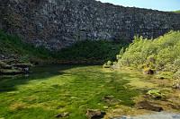 Am grünen Pool  Wir genießen die annähernde Windstille und einen der wenigen, richtigen Sonnentage unserer Reise. Nach nur 25 km biegen wir auf die Straße 85 ab, von der man entlang eines Besucherzentrums zu einem Parkplatz inmitten hellen Grüns gelangt. Wir folgen einem Hinweisschild und gelangen auf einem schön angelegten Waldlehrpfad zu einer Aussichtsplattform am Botnstjörn, einem im Sonnenlicht grün strahlenden Teich direkt an der halbrunden Schluchtwand. Erst auf dem Rückweg bemerken wir, dass es auch einen breiteren, asphaltierten Zuweg vom Parkplatz hierher gibt, bedauern aber nicht, den schöneren Weg durch das aus Birken, Ebereschen und Weiden gebildete Wäldchens genommen zu haben.