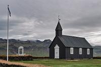 Búðakirkja  ...und die meisten potentiellen Interessenten sitzen noch im Gasthaus beim Frühstück. Die Idee, um 6:30 Uhr aufzustehen und somit bereits kurz vor 7:00 Uhr am Frühstücksbuffet zu sein war brilliant, denn zwanzig Minuten später quoll der Raum über ;-).  Fast gegenüber der Zufahrt zum Wasserfall zweigt eine Straße zur Küste ab. Hier geht es nach Búðir, einem 'Ort', der aus einem Hotel und einer Kirche besteht. Immerhin weht vor der Kirche diesmal tatsächlich eine Islandfahne, die sich aber – wie wir erst am Abend beim Sichten der Tagesausbeute feststellen – just im Moment des Auslösens um den Mast gewickelt hat.