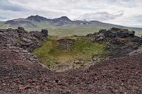 Gipfelstürmer &ast; in  Maren hat es leichtfüßig bis nach oben geschafft und vermeidet es, ebenso wie andere Besucher, sich allzu dicht dem unbefestigten Kraterrand zu nähern, denn zumindest die seeseitige Hälfte besteht aus brauner, kleinkieseliger Vulkanasche, auf der man leicht ins Rutschen kommen kann. Landeinwärts dominieren karstigere Lavafelsen, die einen grünen Kraterboden umschließen und über die man die weiße Gletscherspitze des Snæfellsjökull hinter nähergelegenen Vulkanhügeln erkennt.