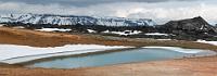 Schwarz, weiß, rot und grün  Etwas oberhalb des Pools, immer noch auf der flach auslaufenden, gelbbraunen Flanke des Leirhnjúkur, bildet ein grünschimmernder Schmelzwassersee mit seinem Schneerand, dem kleinen, aus der schwarzen Ebene aufragenden Vulkankrater und der schneebedeckten Bergkette im Hintergrund ein unwiderstehliches Motiv. Wir folgen dem Bohlenweg noch ein Stück weit, passieren den kleinen Krater und befinden uns in einer von wüsten Lavaauffaltungen gebildeten Landschaft, aus deren Spalten und Rissen überall Dampfschwaden ausgasen.