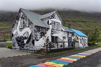 Seyðisfjörður  Die Straße endet direkt im Ortskern und wir stellen unseren Duster auf einem großzügigen Parkplatz neben der Einmündung auf die Ortsstraße ab. Gleich gegenüber liegt ein kleiner See, an dessen linkem Ufer entlang ein bemerkenswerter Fußweg zwischen Hotel, Andenkenshops und Apotheke auf eine hübsche Holzkirche zuführt. Nahezu alle Häuser haben eine Wellblechfassade – bei dem Klima verständlich – von denen die große Mehrheit aber farbenfroh verschönert ist. Ebenso wie der Regenbogenweg, dessen Gestaltung unseres Wissens nicht auf die alberne LSBTIQ+ Wokeness zurückzuführen ist, sondern einfach mit den Häusern harmonieren und mit dem grauen Wetter kontrastieren soll.