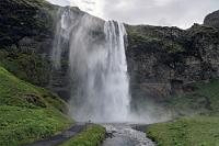 Wat mutt, dat mutt  Mit erheblich gedämpfter Stimmung geht es auf dem gleichen Weg wieder zurück. Meine beiden Nikons samt Objektiven sind von semiprofessioneller Bauart und weitestgehend wasserfest. Der heißgeliebte SONY Camcorder hingegen, den wir völlig unzeitgemäß immer noch der Videofilmerei mit Fotokameras vorziehen, ist als reines Consumerprodukt bei Weitem nicht so hart im Nehmen. Alle Versuche, das Gerät durch Ein- und Ausschalten wiederzubeleben scheitern und ich setze eine kleine Hoffnung auf den Tausch der Batterie, wenn wir wieder beim Auto sind.  Der 61 m hohe Seljalandsfoss bietet beim Queren der kleinen Flußbrücke aber nochmals so einen beeindruckenden Anblick, dass er trotz unserer Stimmung doch noch eine Aufnahme erfordert.